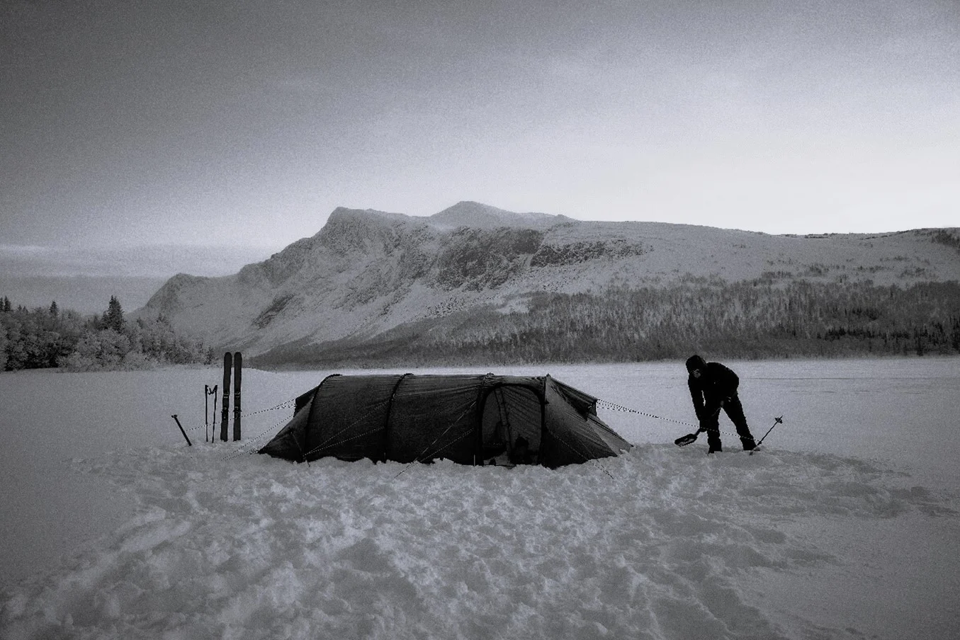 A man shovels snow outside his tent in a snowy landscape.